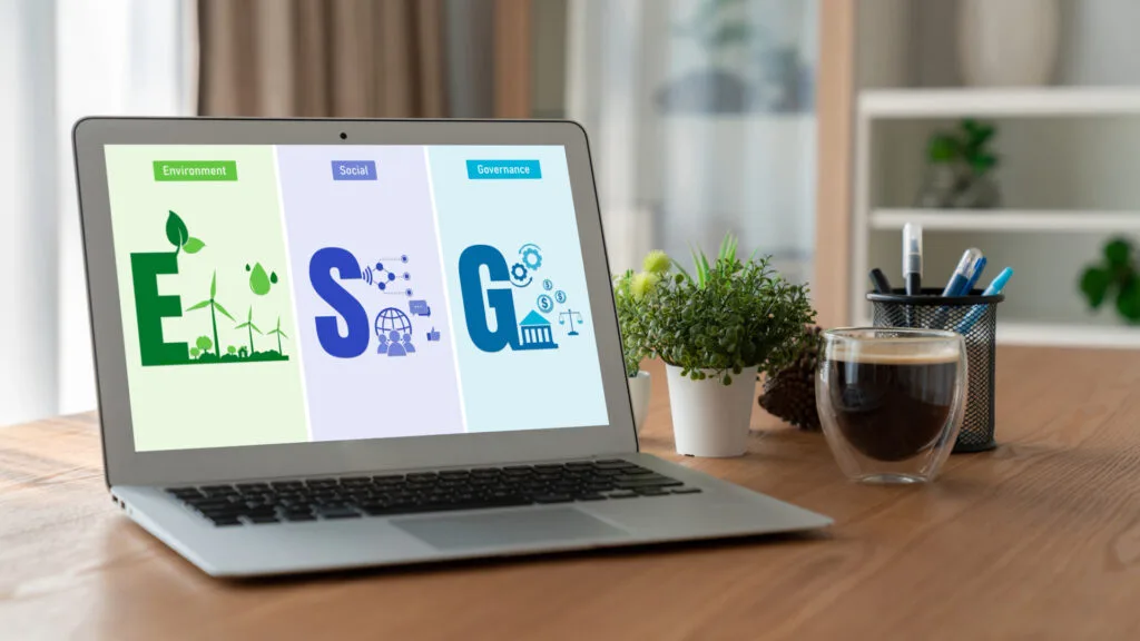 Un laptop che visualizza le categorie ESG sullo schermo: "E" per Ambiente con turbine eoliche e vegetazione, "S" per Sociale con icone di persone e comunicazione, e "G" per Governance con simboli di istituzioni e bilance. Il laptop è posizionato su una scrivania di legno accanto a una tazza di caffè, una pianta in vaso e un portapenne. Lo sfondo presenta scaffali con piante e decorazioni.  